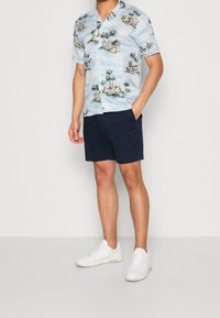 Chemise hawaïenne à manches courtes de couleur bleu clair avec un imprimé de palmiers et de planches de surf, assortie à un short marine foncé et des baskets blanches.