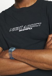 Černé bavlněné tričko s nápisem "NIGHT ADDICT DYSTOPIA" v bílé barvě. Kulatý výstřih, krátké rukávy, nošeno s modrými hodinkami na zápěstí.