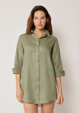 Frau mit langen braunen Haaren, die ein hellolivgrünes Hemdblusenkleid mit hochgekrempelten Ärmeln trägt und vor einem schlichten Hintergrund steht.