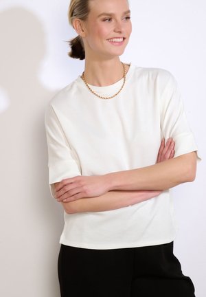 Mujer sonriente con los brazos cruzados, vistiendo una camiseta blanca de manga corta, pantalones negros y un collar de cadena dorada, sobre un fondo liso.