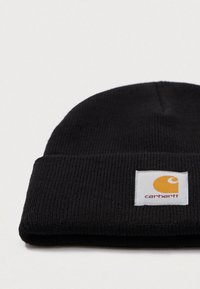 Bonnet en tricot noir à côtes avec un revers, affichant une étiquette carrée blanche portant un logo Carhartt jaune et un texte rouge.