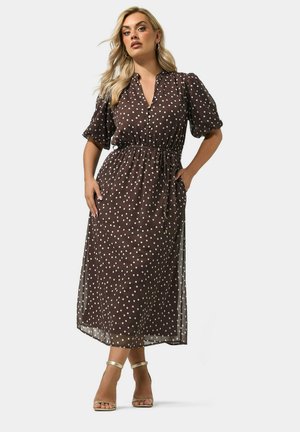 Femme blonde portant une robe midi marron à pois blancs, manches bouffantes, taille cintrée et talons à bride dorés, posant avec une main dans la poche.