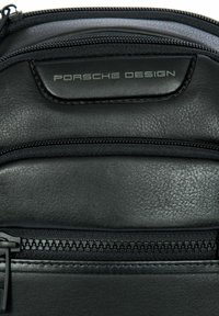 Primo piano di una borsa in pelle nera con più cerniere e un'etichetta con il logo "Porsche Design" sul fronte.