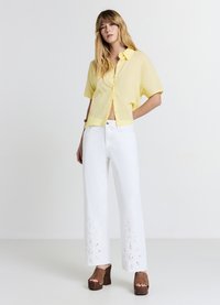 Chemise jaune à manches courtes avec des boutons et une coupe courte, associée à un jean blanc à jambes larges orné de broderies florales sur l'ourlet. Sandales compensées marron.