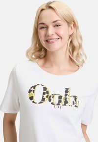T-shirt blanc à manches courtes avec un design graphique portant "Ooh la la !" en vert, imprimé léopard noir et accents en cursive. Matière en coton doux.
