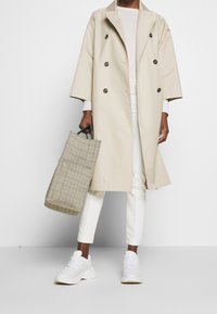 Beige dubbelzijdige trenchcoat met brede mouwen, gedragen over een lichte trui en witte broek. Gestructureerde geruite handtas en witte sneakers.
