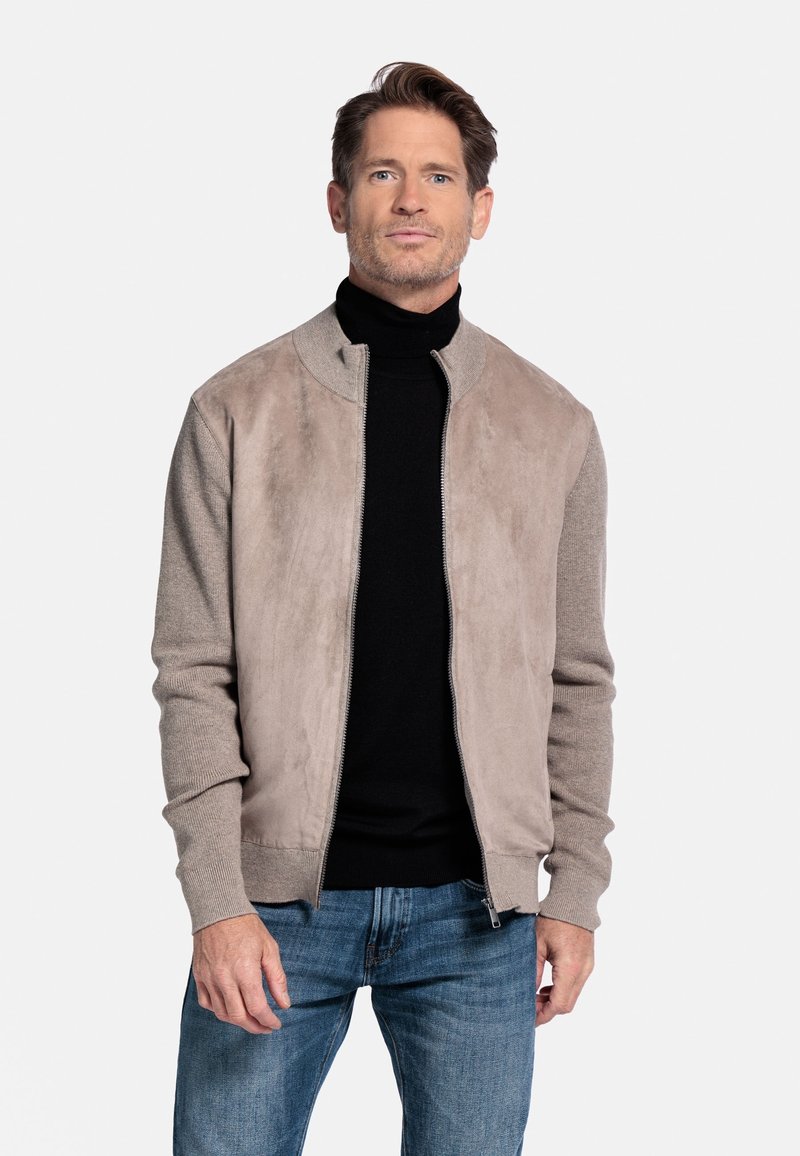 Lichtbeige bomberjack met een zachte suède voorkant en gebreide mouwen. Draag het over een zwarte turtleneck, gecombineerd met blauwe spijkerbroeken.