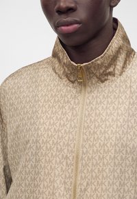 Personne portant une veste beige avec un motif répétitif du logo "MK" et une fermeture éclair dorée, montrée du cou à la poitrine supérieure.