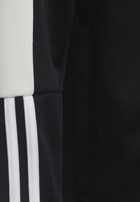 Chaqueta deportiva negra con franjas y paneles blancos, tejido texturizado y un bolsillo con cremallera, destacando un diseño moderno.
