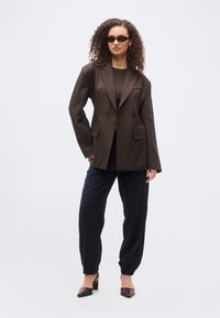 Γυναίκα με σγουρά μαλλιά που φοράει σκούρο, φαρδύ σακάκι, μαύρο τοπ, μπλε σκούρο παντελόνι jogger, μαύρες γόβες και λεπτά γυαλιά ηλίου, στέκεται μπροστά σε λευκό φόντο.