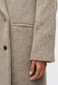 Beżowy wełniany płaszcz o teksturowanej, włochatej powierzchni, z pojedynczym czarnym guzikiem, boczną kieszenią i luźnym rękawem.