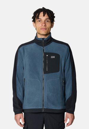 Junger Mann steht und trägt eine blau-schwarze Fleecejacke mit Reißverschluss und Reißverschlusstaschen an den Seiten und an der Brust vor einem einfarbigen Hintergrund.