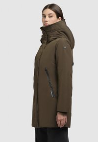 Veste isolée marron avec un col haut et une capuche, dotée de poches zippées et d'une patch logo sur l'épaule. Tissu lisse et durable.