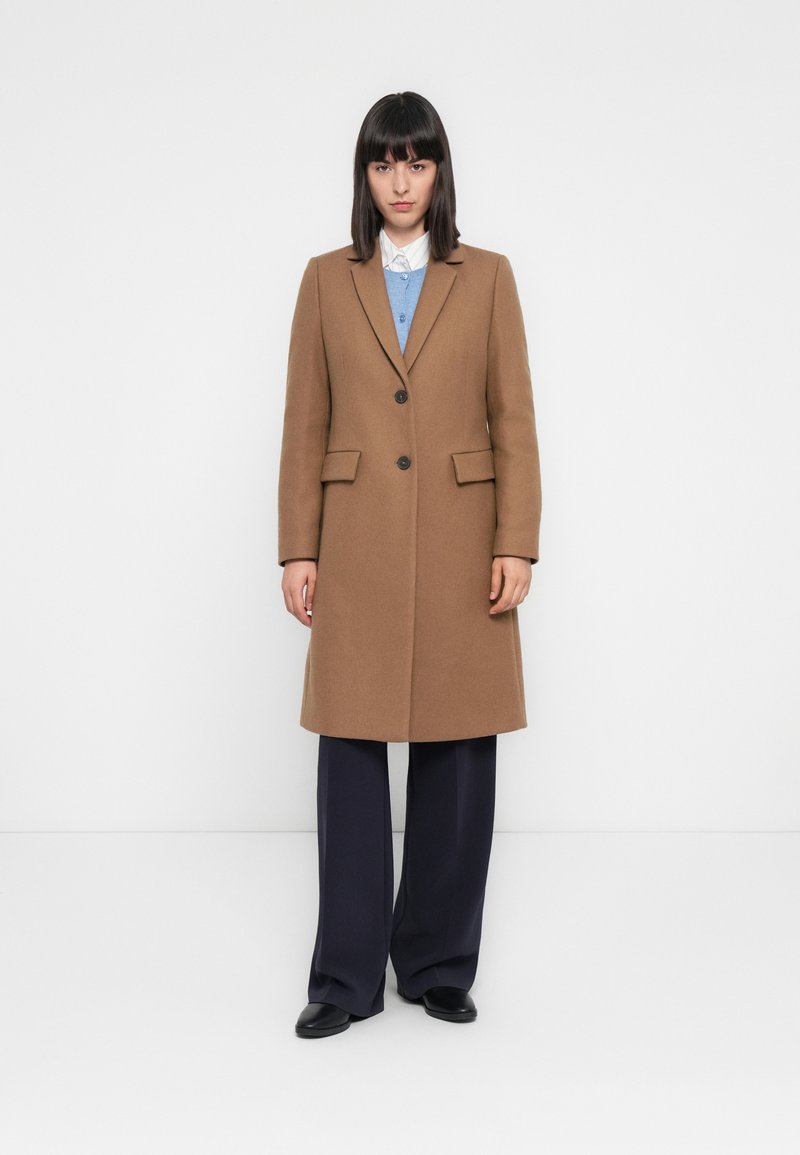 Femme portant un manteau marron arrive-genoux par-dessus une chemise blanche et un pull bleu, avec un pantalon large noir et des chaussures noires, se tenant contre un mur blanc.
