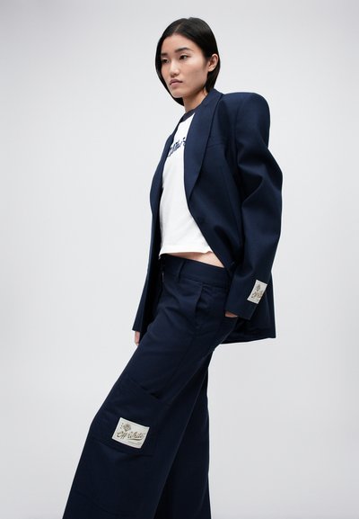Námořnický modrý blazer a kalhoty s širokými nohavicemi z pevných materiálů, pod kterým je bílý tričko s logem a detailní nášivky na kalhotách.