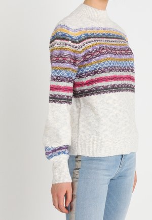 Frau trägt einen hellgrauen Pullover mit mehrfarbigen horizontalen Streifen über Brust und Ärmeln, kombiniert mit hellblauen Jeans mit seitlichen Streifen.