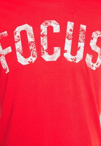 Camiseta de algodón roja con grandes letras blancas en bloque que dicen "FOCUS", con un patrón floral dentro de las letras. Diseño de mangas cortas y cuello redondo.