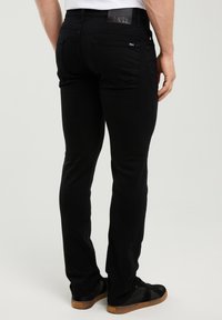 Mann trägt schwarze schlanke Jeans und schwarze Turnschuhe mit braunen Sohlen, dargestellt von hinten vor einem schlichten hellen Hintergrund.