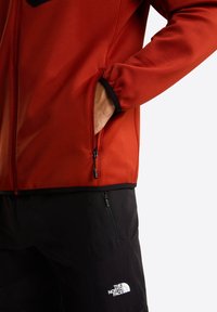 Rode fleecejas met een ritszak, zwarte manchetten en een contrasterende zwarte borstzak. Gecombineerd met zwarte shorts met een logo.