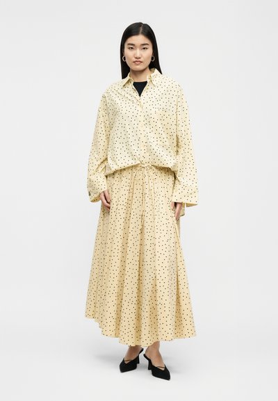 Γυναίκα που φοράει κρεμ μπλούζα με κουμπιά και μακριά μανίκια, καθώς και μακριά φούστα με μαύρες βούλες, σετ με μαύρα μυτερά παπούτσια.