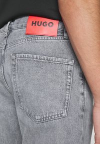 Lähikuva harmaista farkuista, joissa on takatasku ja pieni metallinen Hugo-tunnus sekä punainen Hugo-brändilogo vyötäröllä, käytetty mustassa paidassa olevan henkilön päällä.