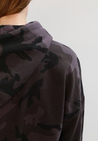 Sudadera con capucha de diseño en camuflaje púrpura y negro, con una capucha amplia y tejido suave, mostrando un primer plano del diseño y la textura en la parte posterior.