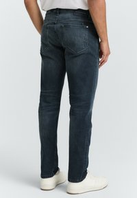 Jeans de mezclilla azul oscuro con un corte ajustado, que presentan bolsillos en la parte posterior y un detalle mínimo. Se llevan con zapatillas blancas, mostrando un aspecto limpio y casual.