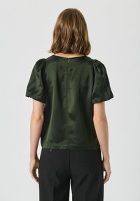 Blusa di raso verde scuro con maniche corte a sbuffo, scollatura rotonda e una cerniera discreta sul retro. Texture liscia e lucida, vestibilità classica.