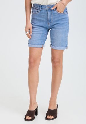 Donna con shorts di jeans azzurri arrotolati e mules nere aperte davanti con tacco, in piedi su uno sfondo chiaro e uniforme.
