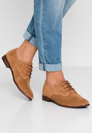 Light brown suede lace-up shoes worn with rolled-up light blue denim jeans, shown on a white floor with a plain background.