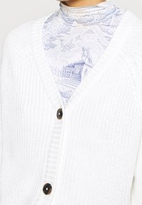 Cárdigan blanco de punto con escote en V, que presenta una textura acanalada y dos grandes botones oscuros. Debajo lleva una blusa de color azul claro con un estampado.