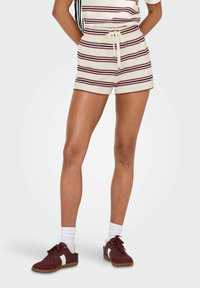 Shorts a righe crema con linee rosse e blu navy, dotati di una cintura elastica con lacci. Abbinati a sneakers bordeaux e calze bianche.