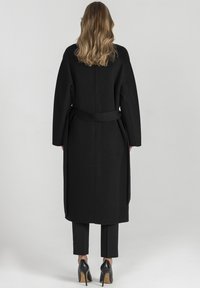 Czarny wełniany płaszcz z szerokimi rękawami, paskiem w talii i prostym krojem. Poniżej płaszcza widać czarne spodnie i wysokie obcasy.
