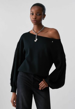 Frau trägt schwarzen schulterfreien Pullover mit strukturierten Ballonärmeln, schwarze Hose, silberne Kette mit Herzanhänger und silberne Ohrringe.