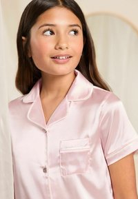 Camicia da pigiama di seta rosa chiaro con colletto a punta, maniche corte e taschino sul petto con dettagli di cucitura sottili. Chiusura con bottoni al centro.