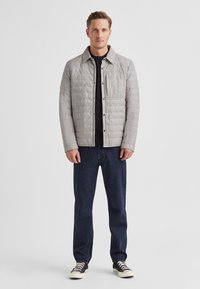 Hellgraue gesteppte Jacke mit Kragen, Fronttasche und Druckknöpfen, kombiniert mit dunklen Jeans und schwarzen Sneakers.