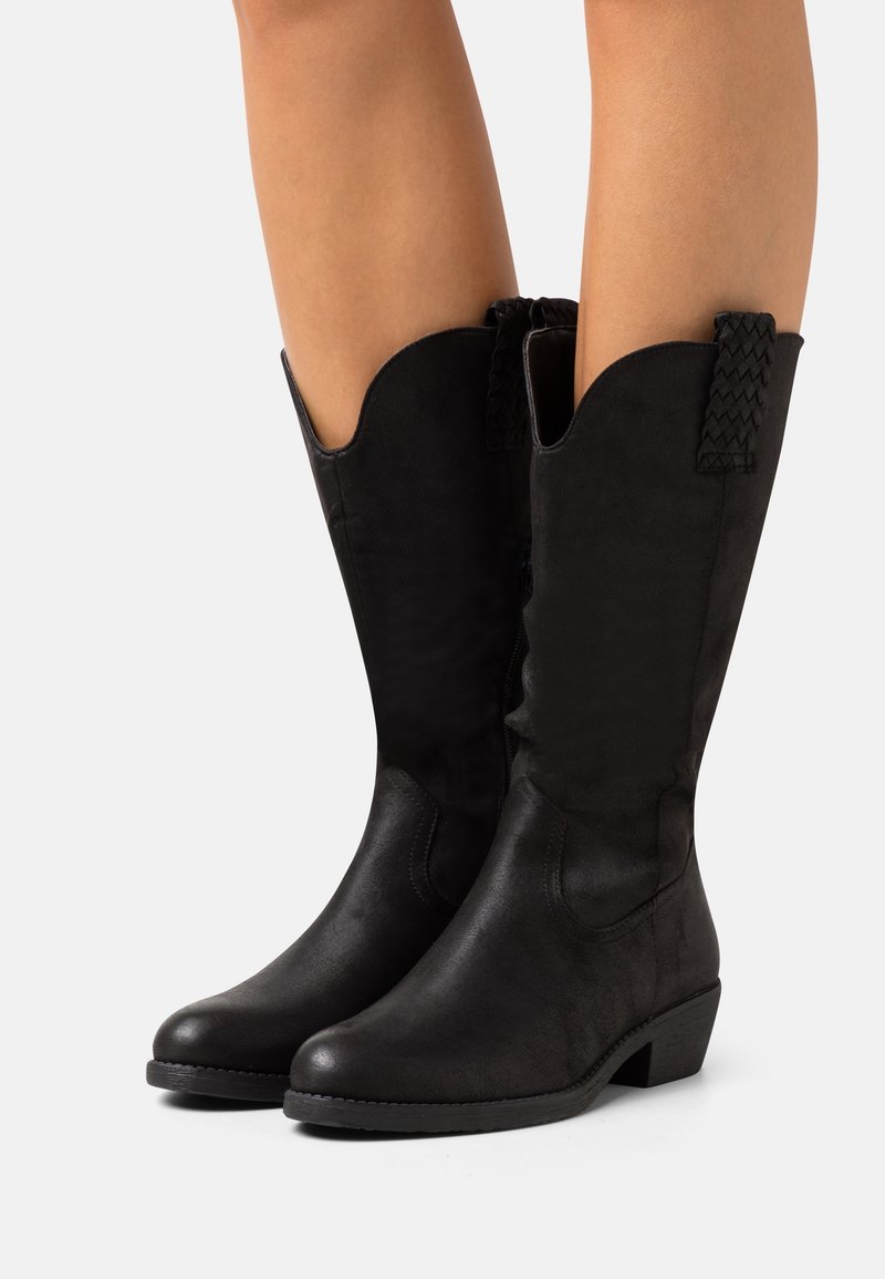 Paire de bottes en cuir noires mi-mollets avec talons bas et languettes, portées sur des jambes nues sur fond blanc.