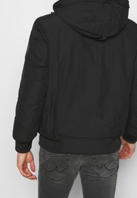 Schwarze gepolsterte Jacke mit Kapuze, ausgestattet mit einer glatten Textur, elastischen Bündchen und einem geraden Saum. Detail am linken Ärmel. Kombiniert mit grauen Jeans.