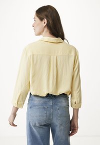 Camicia gialla chiaro a maniche lunghe con polsini arrotolabili, vestibilità ampia, dettaglio con piega sul retro, abbinata a jeans in denim di lavaggio medio con tasche posteriori.