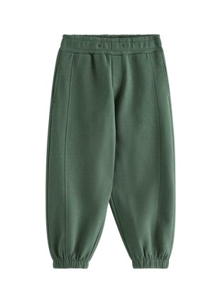 Et ungt barn smiler med hænderne på hoften iført en blå strikhue, denimjakke, brun skjorte med gule lastbiler, grønne bukser og blå sneakers.