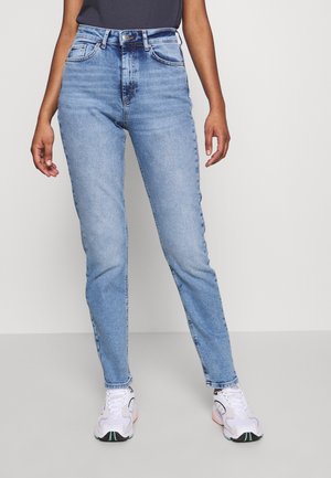 Personne portant un jean taille haute bleu clair, un haut noir et des baskets blanches, debout devant un fond gris uni.
