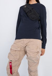 Osoba w granatowej koszuli z długim rękawem, beżowych spodniach cargo z klapkami oraz czarnej torbie na ramię z zamkami i małą naszywką z logo.