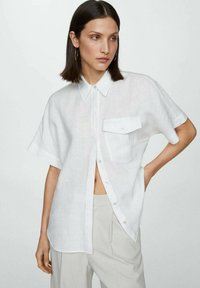 Camicia bianca a maniche corte con bottoni in tessuto leggero, caratterizzata da un colletto a punta e una tasca sul petto sinistro con pattina.