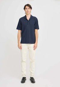 Marineblå kortærmet skjorte med knapper, tekstureret stof, båret med cremede jeans og sorte sko. Enkelt krave og frontlukning med knapper.