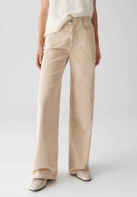 Beige Cordhosen mit weitem Bein, strukturiertem Stoff, Fronttaschen und einem Knopfverschluss am Bund. Kombiniert mit einem hellen Oberteil und Schuhen.