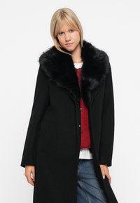 Cappotto nero con colletto in pelliccia sintetica, maniche lunghe e tasche laterali, sovrapposto a un maglione rosso a maglia e jeans blu. Texture liscia.