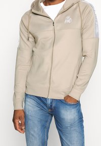 Beige zip hoodie med dragsko, med vit logobroderi och randiga detaljer på axlarna. Mjuk tyg, avslappnad passform.