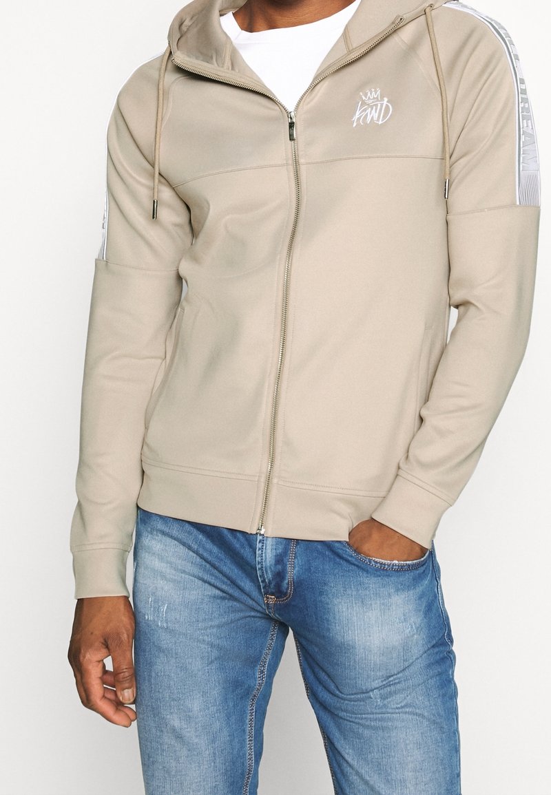Beige zip hoodie med dragsko, med vit logobroderi och randiga detaljer på axlarna. Mjuk tyg, avslappnad passform.