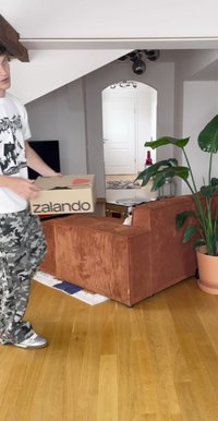 Kartónová krabice s logem "zalando", držena nad dřevěnou podlahou. V pozadí hnědá kordurová pohovka a vysoká zelená rostlina v květináči.