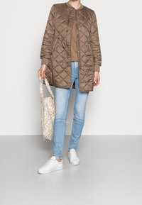 Pikowana brązowa długa kurtka z okrągłym kołnierzem, zestawiona z beżową bluzką, jasnoniebieskimi dżinsami i białymi trampkami. Trzyma wzorzysty torbę na ramię.